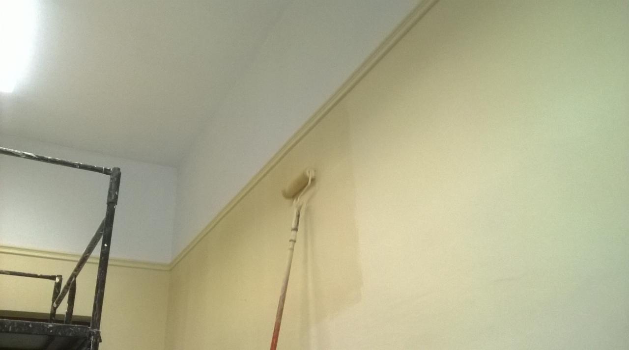 Świeżo pomalowana ściana w odcieniach beżu z widoczną linią odcięcia koloru przy suficie, z wałkiem malarskim i fragmentem rusztowania w kadrze.
