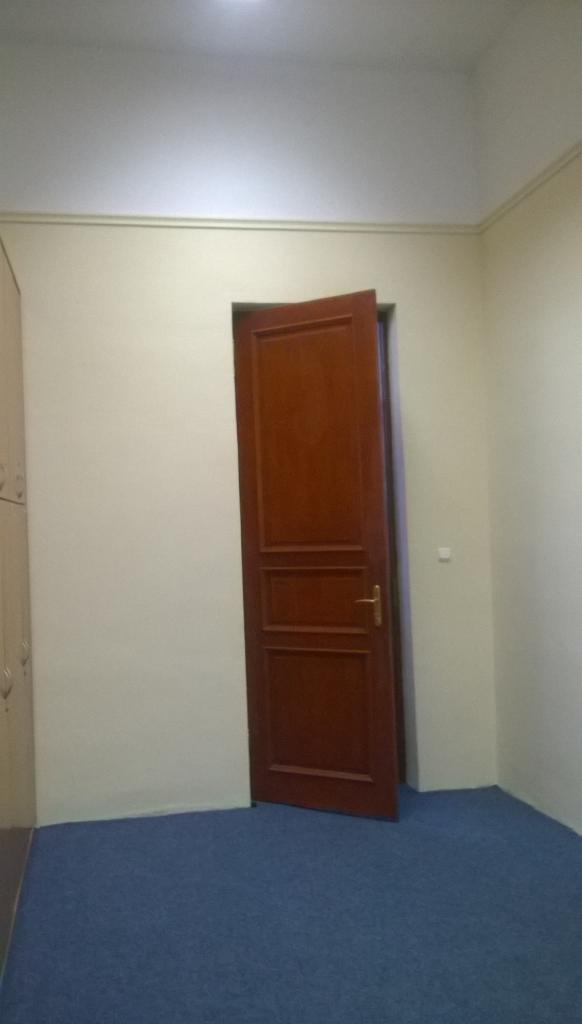 Pokój z beżowymi ścianami, niebieską wykładziną i częściowo otwartymi, brązowymi drzwiami z trzema panelami, oświetlony górnym światłem.