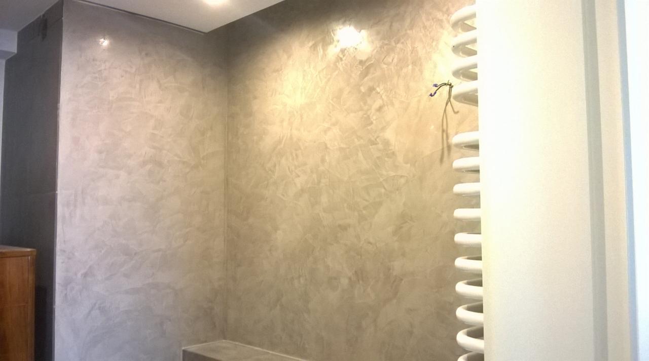 Ściana w łazience wykończona dekoracyjną, szarą gładzią w stylu weneckim, z widocznym grzejnikiem drabinkowym i fragmentem szafki.