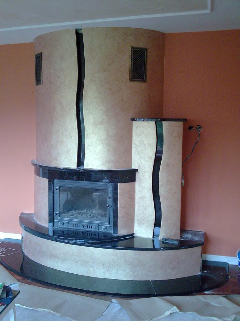 Wykończony kominek z obudową w odcieniach beżu i czerni, z wyraźną fakturą na ścianach i elementach dekoracyjnych, widoczne kratki wentylacyjne i czarny blat.