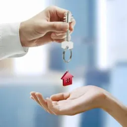 Przekazanie kluczy z czerwoną zawieszką w kształcie domu z jednej dłoni do drugiej na rozmytym, jasnym tle.