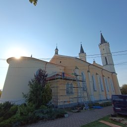 PRZEDSIĘBIORSTWO WIELOBRANŻOWE MARCIN BIENIO - Prace na dachu kościoła w promieniach wschodzącego słońca, widoczne rusztowania i pracownik w niebieskim ubraniu, częściowo zdjęte pokrycie dachu.