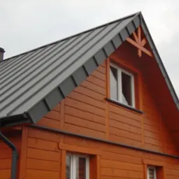 Dom w Suwałkach z nowym, szarym pokryciem dachowym z blachy trapezowej, widoczny komin i drewniana elewacja w odcieniach brązu.