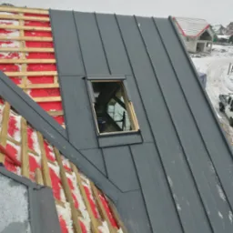 Montaż szarego pokrycia dachowego z blachy na nowym budynku z oknem dachowym, widoczne drewniane łaty i izolacja, częściowo pokryte śniegiem.