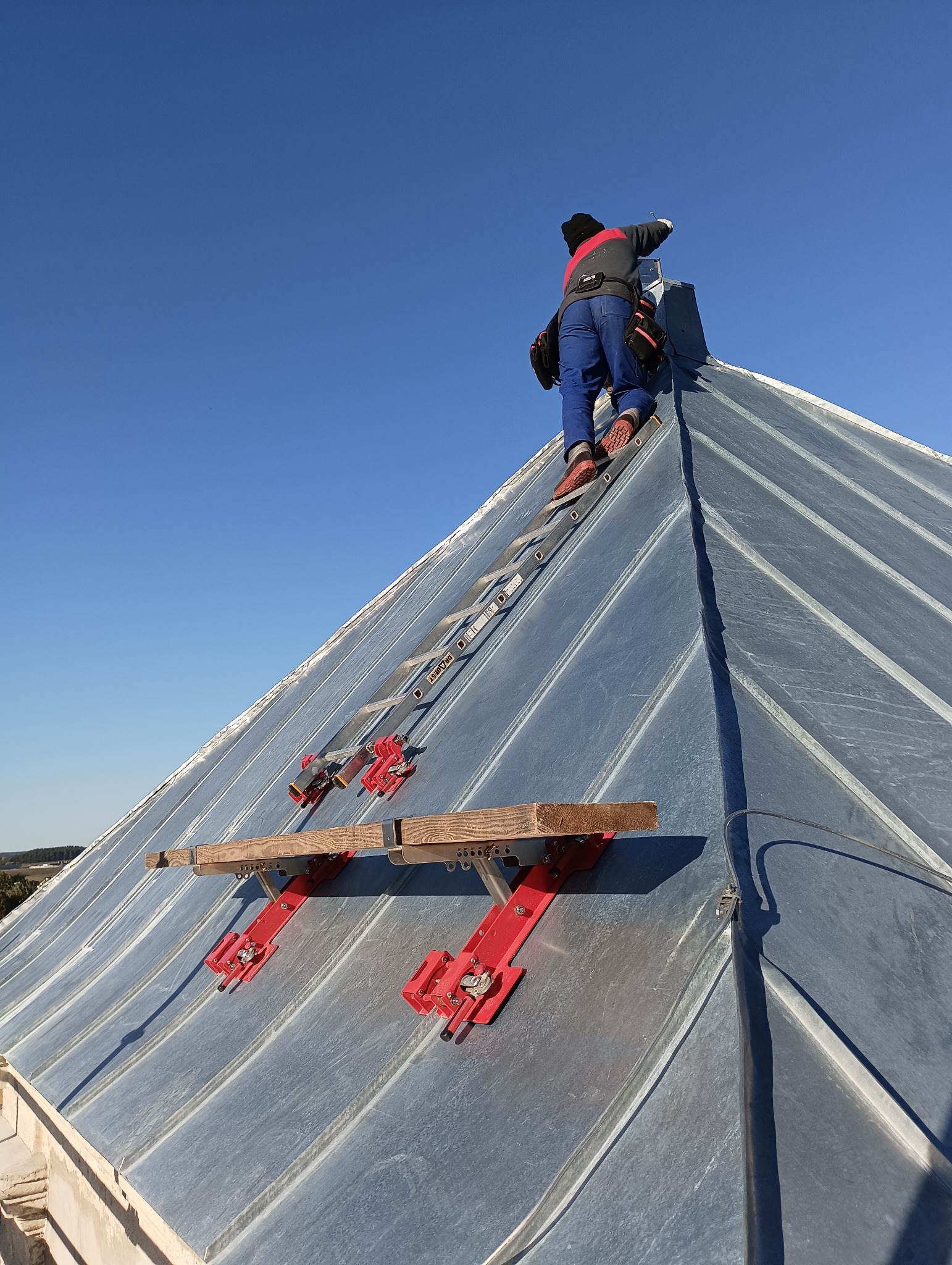 Dekarz na drabinie mocowanej do dachu z blachy trapezowej, wykonujący prace na szczycie dachu w słoneczny dzień.