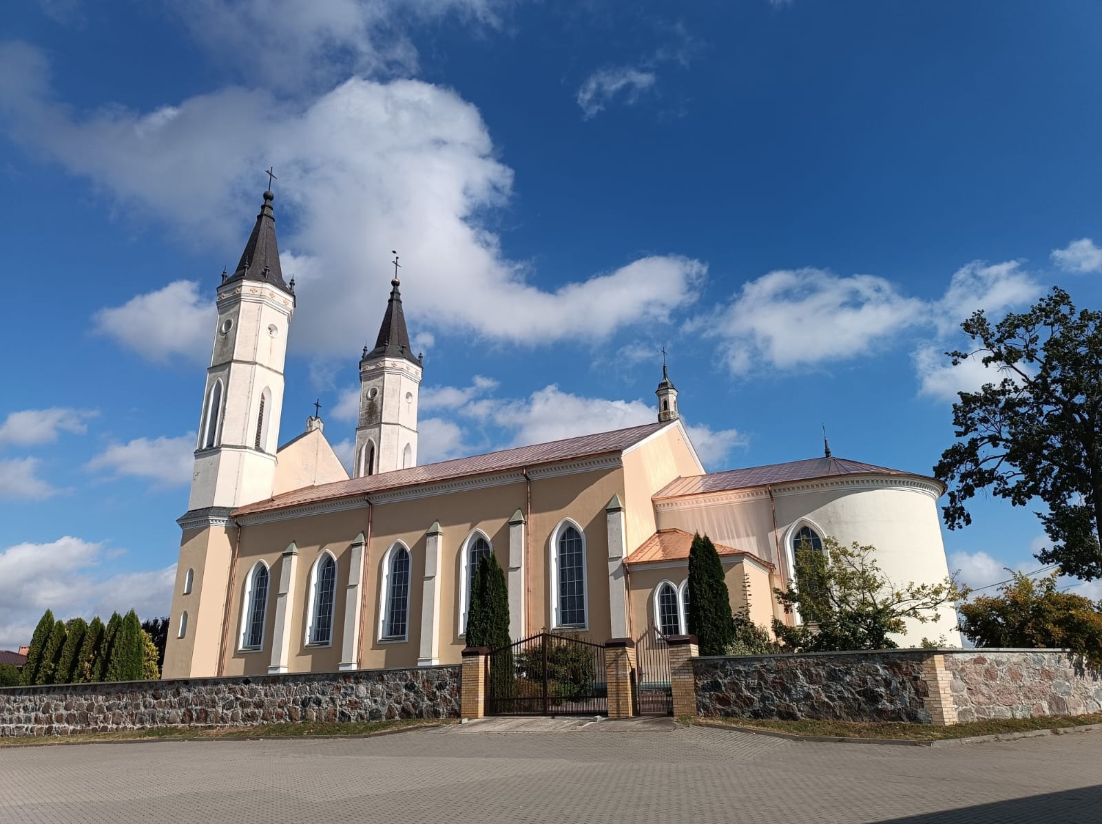 Kościół z dwiema wieżami i miedzianym dachem w słoneczny dzień, widoczne detale architektoniczne i kamienne ogrodzenie.