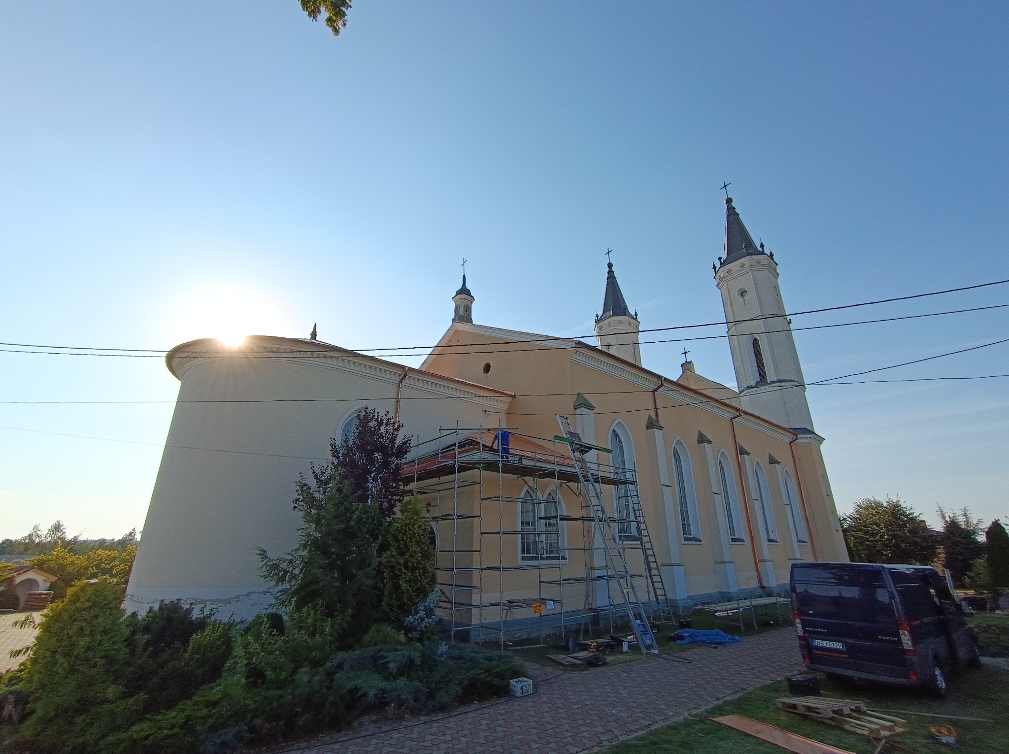 Prace na dachu kościoła w promieniach wschodzącego słońca, widoczne rusztowania i pracownik w niebieskim ubraniu, częściowo zdjęte pokrycie dachu.