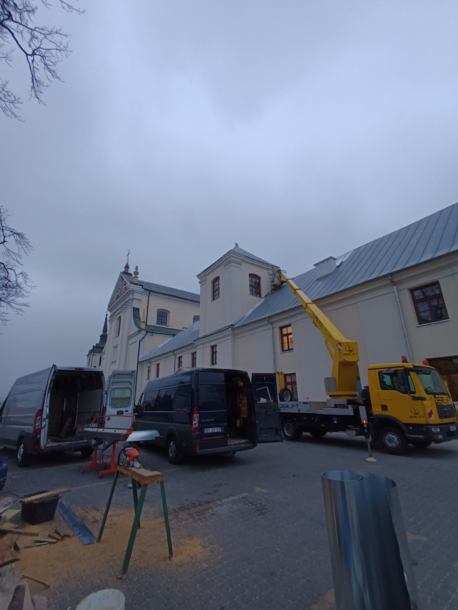 Prace dekarskie przy zabytkowym budynku: żółty podnośnik koszowy przy dachu, otwarte busy, narzędzia do obróbek blacharskich na chodniku.