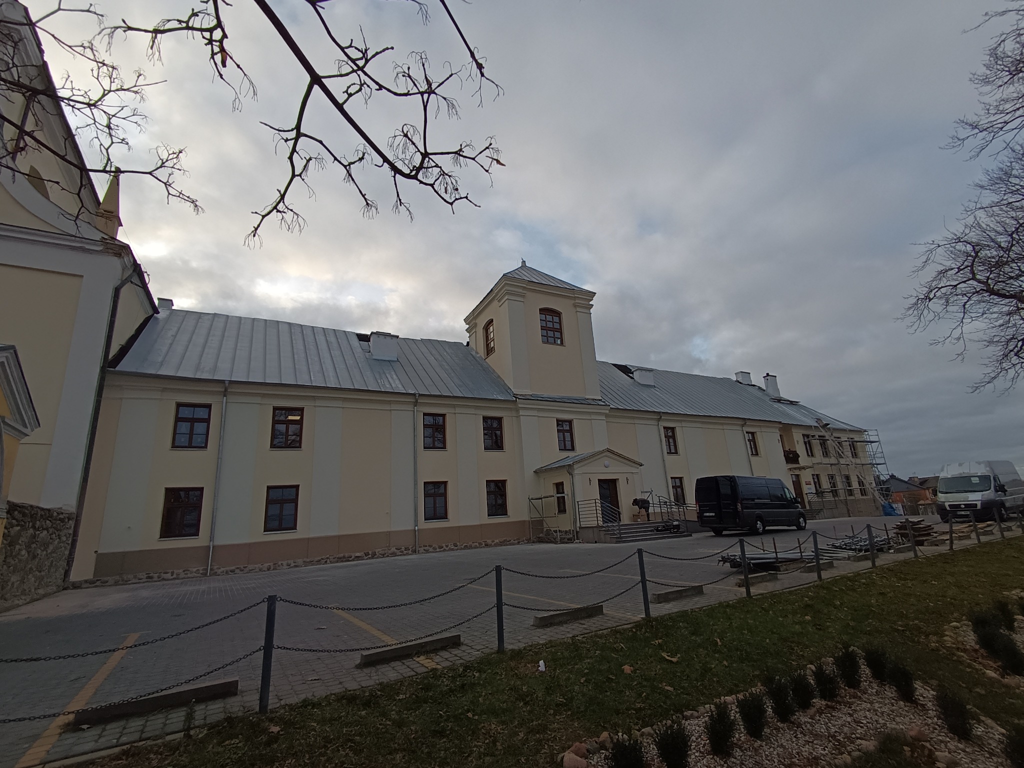 Budynek z jasną elewacją i szarym dachem, widoczne rusztowania i zaparkowane busy, pochmurne niebo w tle.