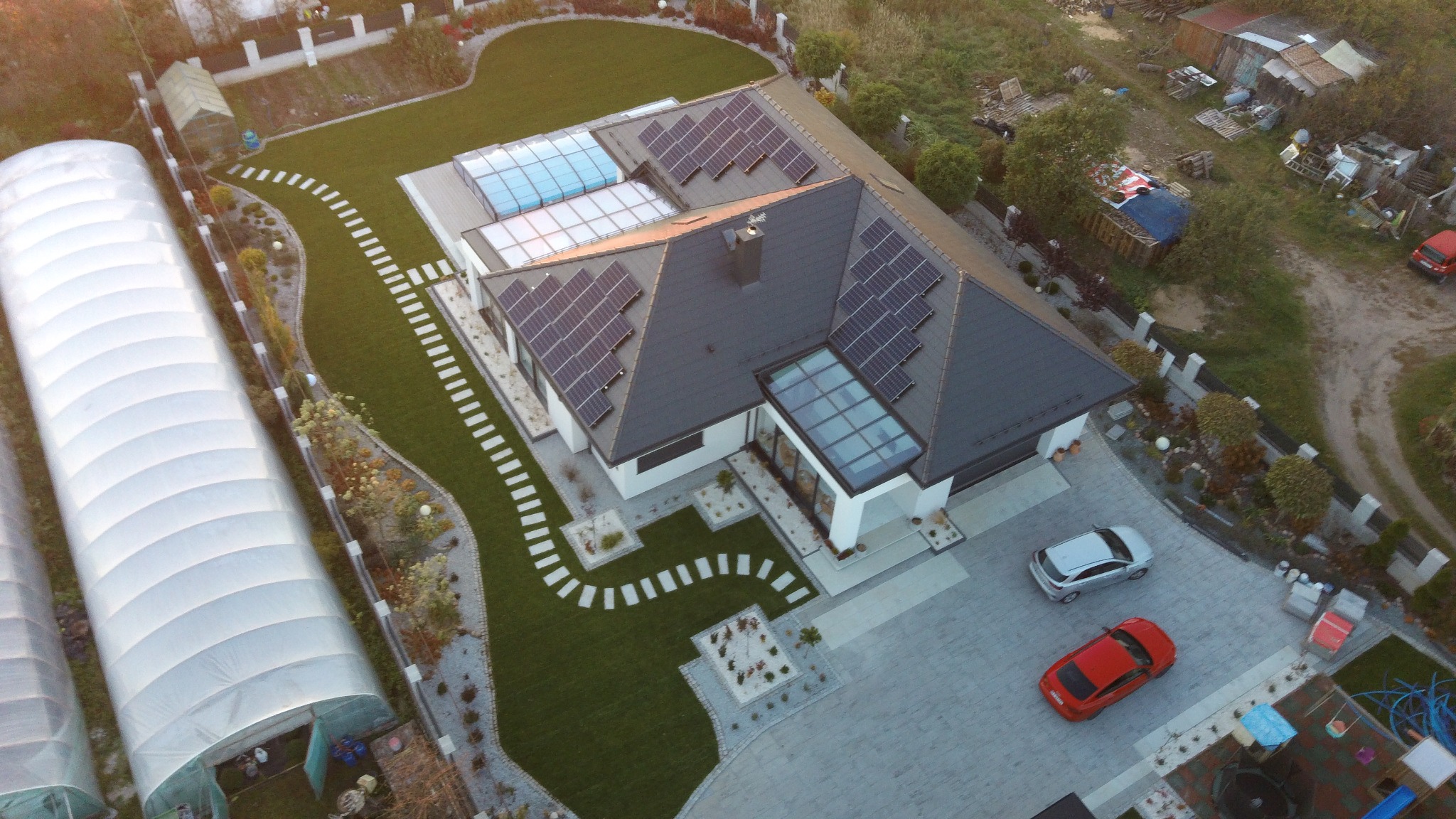 Widok z lotu ptaka na nowoczesny dom z ciemnoszarym dachem, panelami słonecznymi i przeszklonymi fragmentami, otoczony zadbanym ogrodem z chodnikiem z płyt i dwoma samochodami na podjeździe.