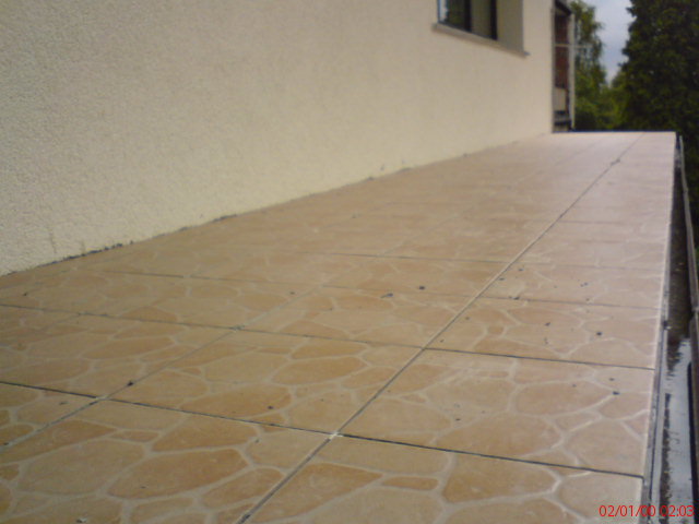 Wykafelkowany taras z beżowymi płytkami o nieregularnym wzorze, widok z boku, jasna elewacja budynku w tle.