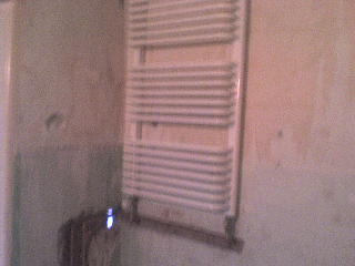 Biały grzejnik łazienkowy drabinkowy zamontowany na surowej, nierównej ścianie z widocznymi śladami po poprzednich pracach. W tle fragment starego, zardzewiałego grzejnika.