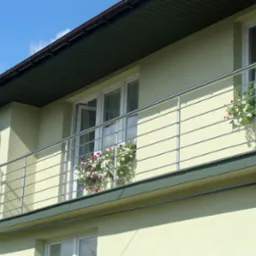 Balkon z nierdzewną balustradą z wypełnieniem z poziomych prętów skręconych w spiralę, donice z kwiatami na balkonie, antena satelitarna na elewacji budynku.