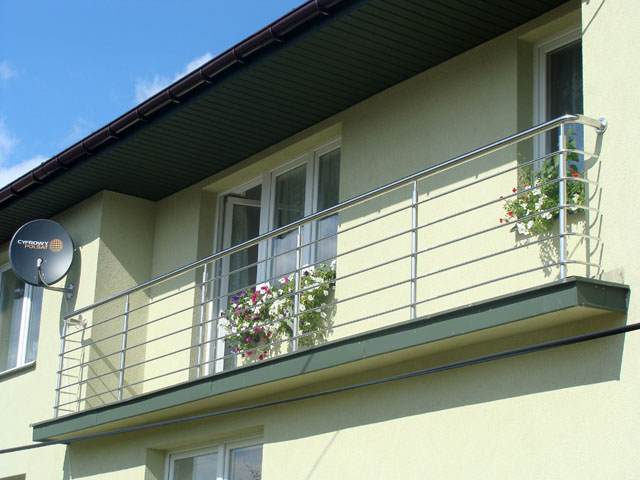 Balkon z nierdzewną balustradą z wypełnieniem z poziomych prętów skręconych w spiralę, donice z kwiatami na balkonie, antena satelitarna na elewacji budynku.