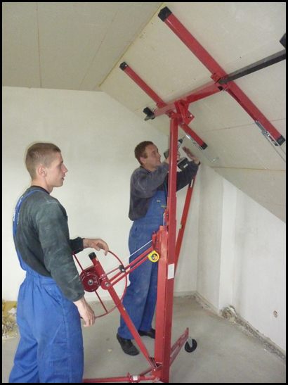 Dwóch monterów w niebieskich kombinezonach instaluje płyty gipsowo-kartonowe na poddaszu za pomocą podnośnika do płyt. Jeden monter przykręca płytę wkrętarką, drugi obsługuje mechanizm podnoszący.