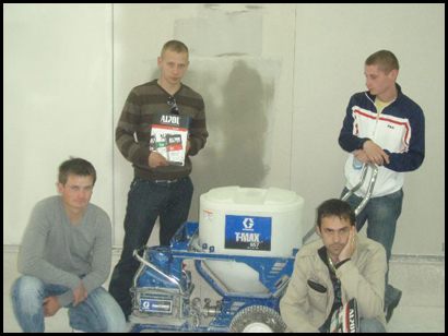Grupa czterech mężczyzn pozuje obok maszyny do natrysku tynku Graco T-MAX 657 na tle ściany z częściowo nałożonym tynkiem. Jeden z mężczyzn trzyma katalog ALJO701.