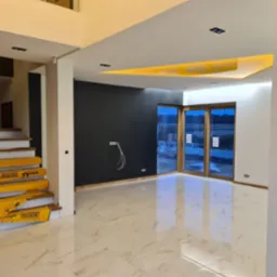 Nowoczesne wnętrze domu w trakcie wykańczania: białe marmurowe płytki podłogowe, czarna ściana z wystającymi przewodami elektrycznymi, schody zabezpieczone ż&oacute;łtą folią ochronną, przesuwne drzwi...