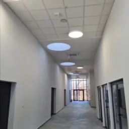 Wnętrze wyremontowanego, jasnego korytarza biurowego w Szastarce z oświetleniem sufitowym i szarą posadzką, widok w kierunku przeszklonego wyjścia.