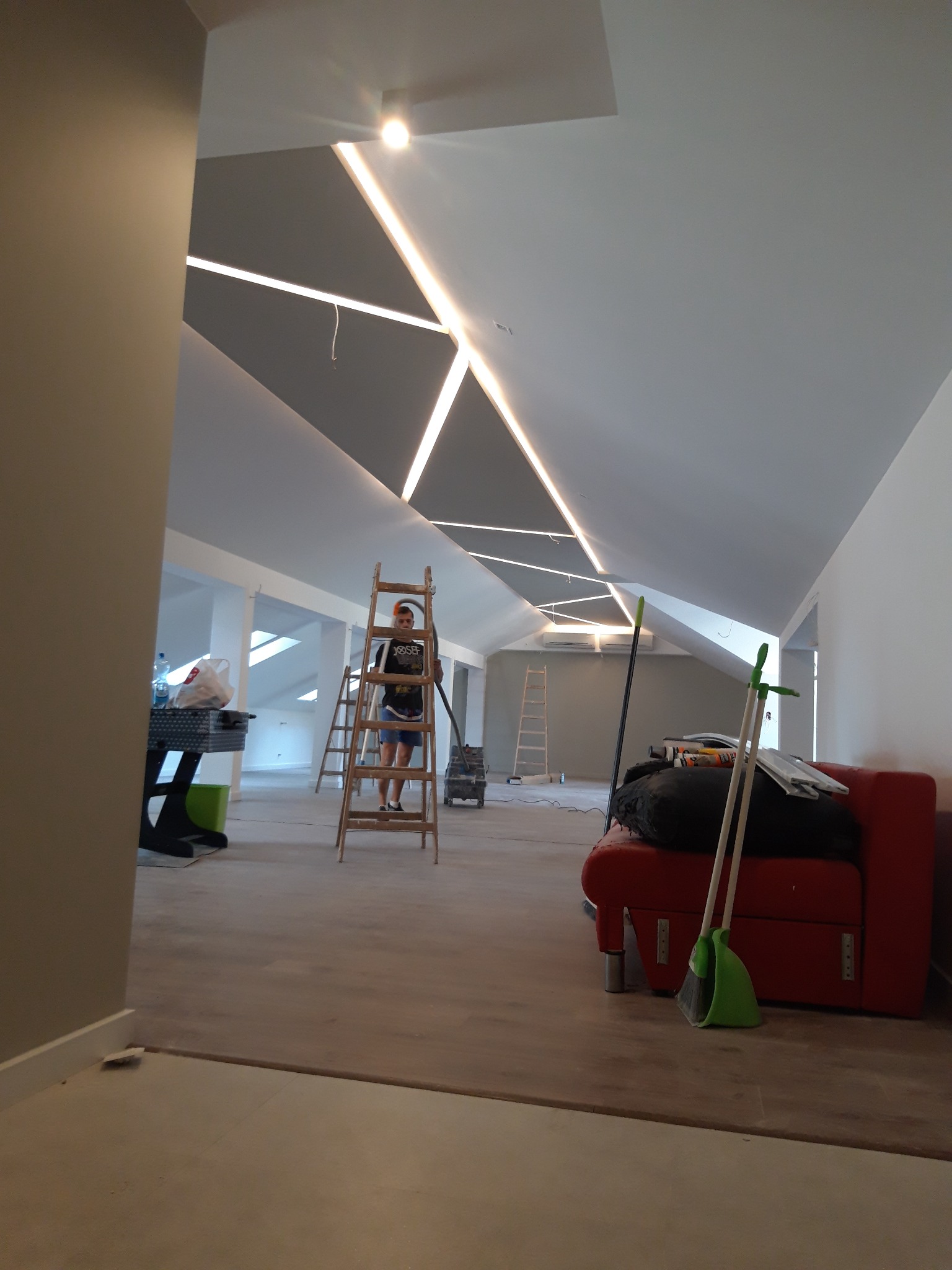 Wnętrze poddasza w trakcie prac wykończeniowych, widoczne oświetlenie LED w suficie, drabiny oraz osoba na drabinie, przygotowująca się do dalszych prac.