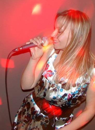 Kobieta z blond włosami w kwiecistej sukience śpiewa do mikrofonu oświetlonego czerwonym światłem.