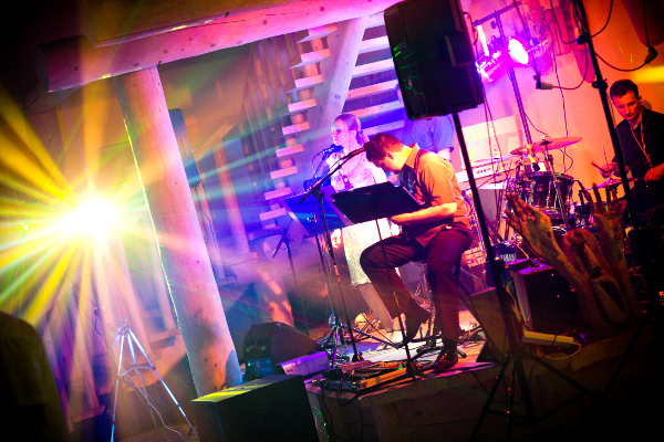 Zespół muzyczny grający koncert na scenie z drewnianymi elementami konstrukcyjnymi w tle, oświetlonej kolorowymi światłami. Widać perkusję, keyboard i wokalistów.