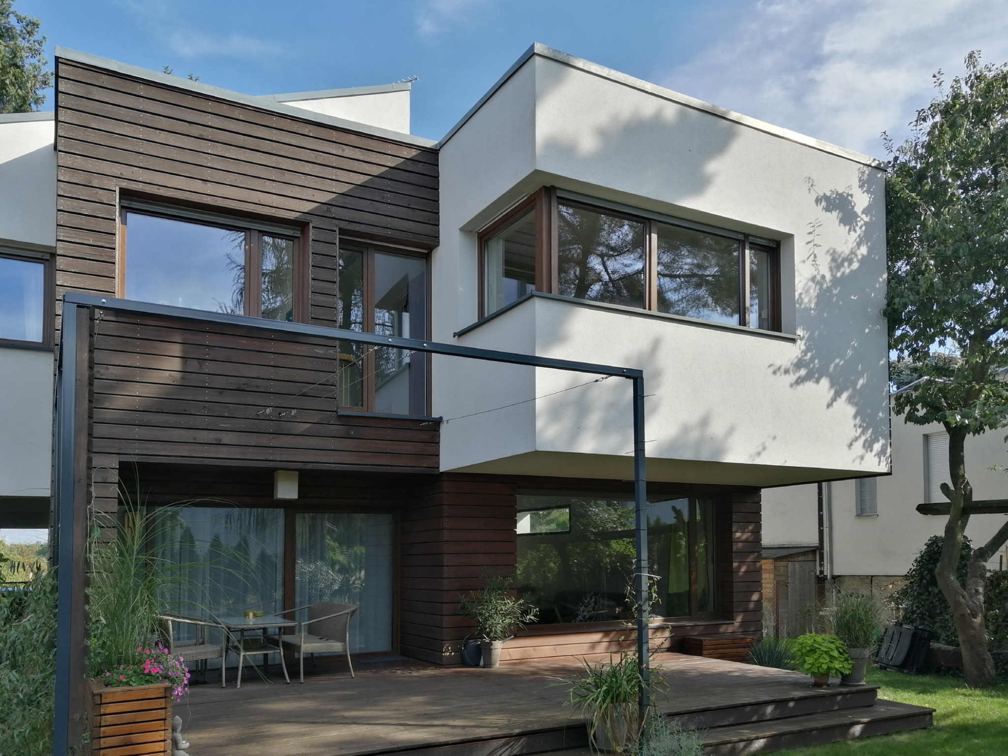 Nowoczesny dom z elementami drewnianej elewacji i tarasem, z widocznymi meblami ogrodowymi i roślinnością