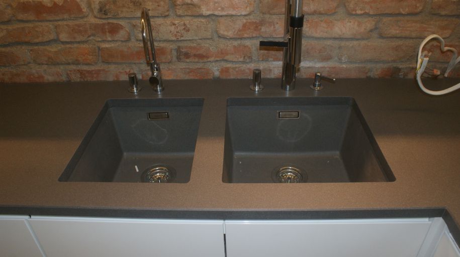 Dwa identyczne, grafitowe zlewozmywaki z bateriami zamontowane w blacie z konglomeratu na tle ściany z czerwonej cegły i białych szafek kuchennych.