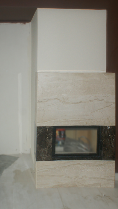 Fragment obudowy kominka z jasnego kamienia naturalnego z ciemnym, marmurkowym obramowaniem w trakcie montażu, widoczne niedokończone ściany i podłoga.