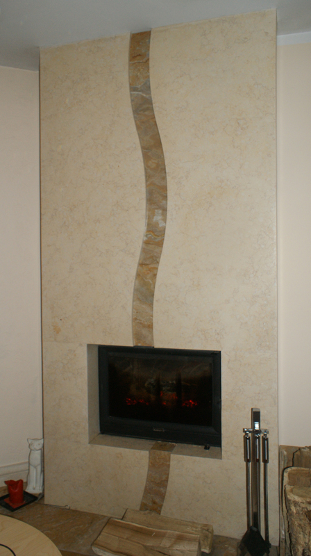 Obudowa kominka wykończona beżowym kamieniem z falistym, dekoracyjnym pasem z jaśniejszego kamienia, widoczne akcesoria kominkowe i drewno.