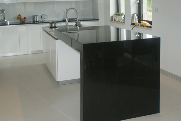 Nowoczesna kuchnia z czarnym, błyszczącym blatem wyspowym, białe szafki i jasna podłoga, widok na okno z roślinami w tle.