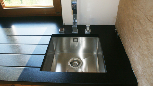 Nowoczesny, prostokątny zlew ze stali nierdzewnej w czarnym, kamiennym blacie kuchennym z frezowanymi rowkami, bateria chromowana, ściana wykończona kamieniem dekoracyjnym.