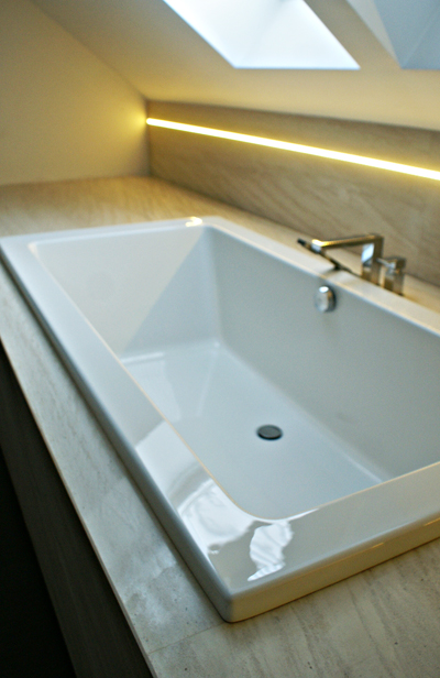 Nowoczesna biała wanna wbudowana w blat z jasnego trawertynu, podświetlenie LED pod sufitem, okno dachowe.