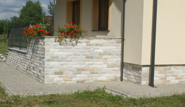 Elewacja domu wykończona kamieniem naturalnym w odcieniach beżu i szarości, uzupełniona rabatą z czerwonymi kwiatami w donicach, widoczna część chodnika z kostki brukowej.