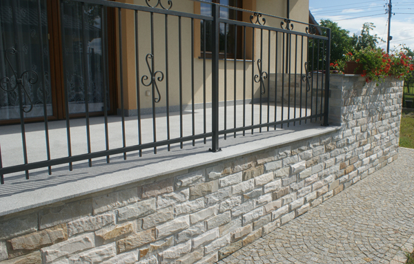 Elewacja budynku wykończona kamieniem naturalnym, z widocznym fragmentem balkonu z czarną, metalową balustradą i ścieżką z kostki brukowej.