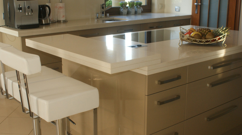 Beżowa wyspa kuchenna z blatem z konglomeratu kwarcytowego, owocami w ozdobnej misie i białym krzesłem barowym.