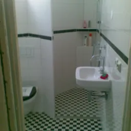 Wnętrze łazienki z toaletą, umywalką i prysznicem, wykończone białymi płytkami z czarnym pasem i podłogą w czarno-białą szachownicę, widziane zza framugi drzwi.