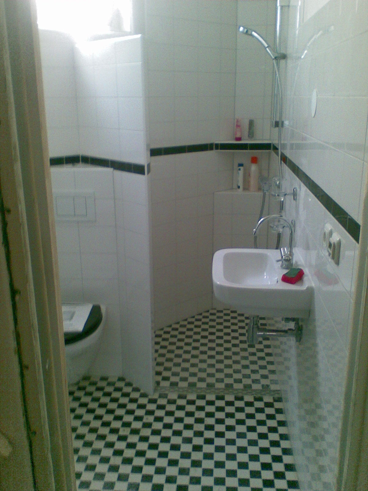 Wnętrze łazienki z toaletą, umywalką i prysznicem, wykończone białymi płytkami z czarnym pasem i podłogą w czarno-białą szachownicę, widziane zza framugi drzwi.