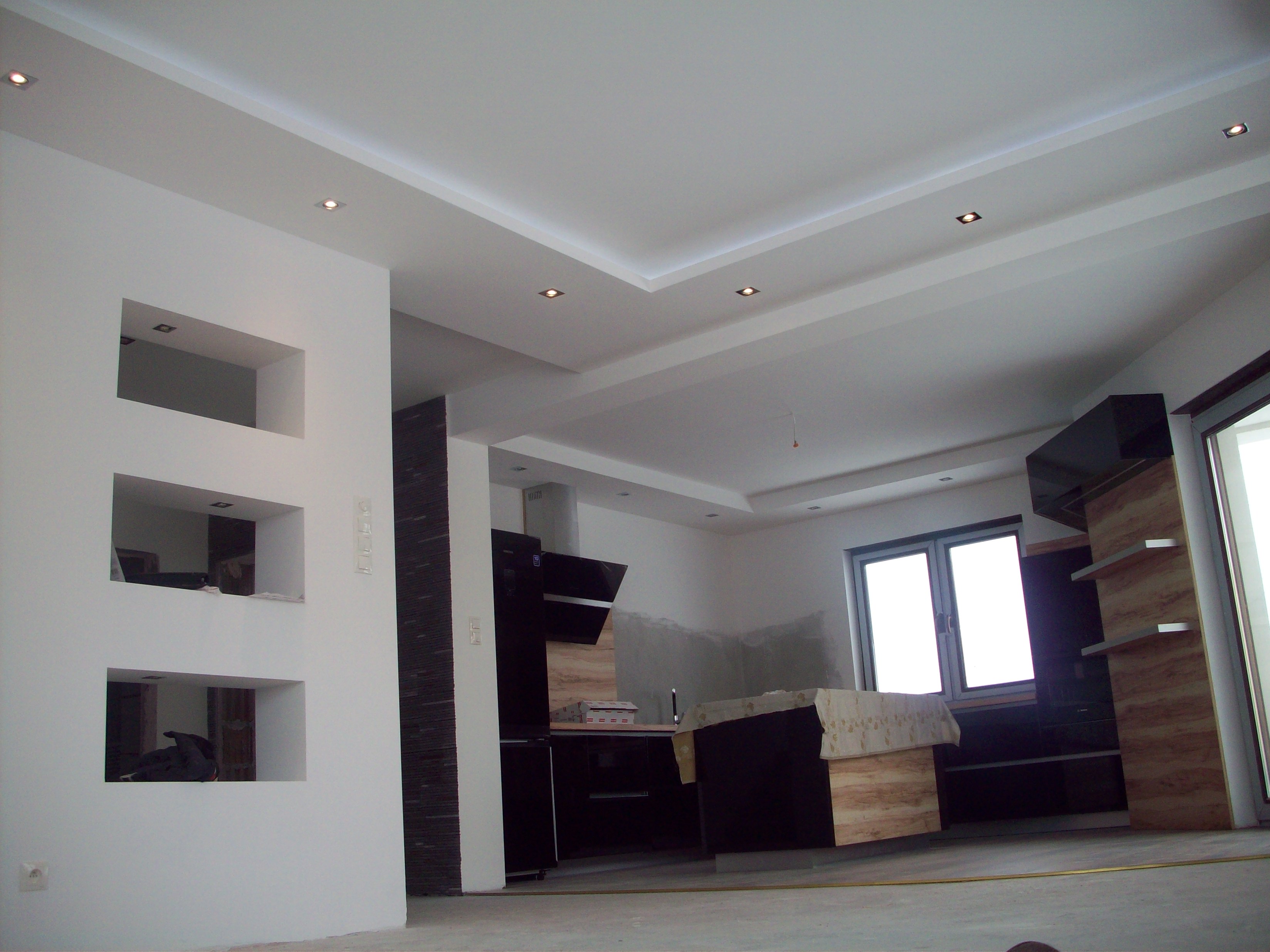 Wnętrze kuchni w trakcie remontu, widoczne czarne szafki, fragmenty ściany z wnękami i oświetlenie sufitowe.