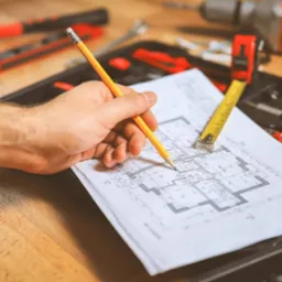 Ręka z ołówkiem nad planem domu, obok zwinięta miara, w tle narzędzia w skrzynce.