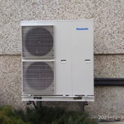 Zewnętrzna jednostka pompy ciepła Panasonic zamontowana na elewacji budynku z szorstkim tynkiem, widoczne rury w izolacji termicznej.