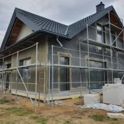 Dom w trakcie ocieplania elewacji: widoczne rusztowania, warstwa izolacyjna, częściowo nałożony tynk, okna zabezpieczone folią, bloczki budowlane i materiały budowlane na ziemi.