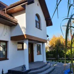 Dwupiętrowy dom z białą elewacją, brązowymi elementami drewnianymi i schodami z szarej kostki brukowej, widok z poziomu gruntu, z elementami roślinności w kadrze.