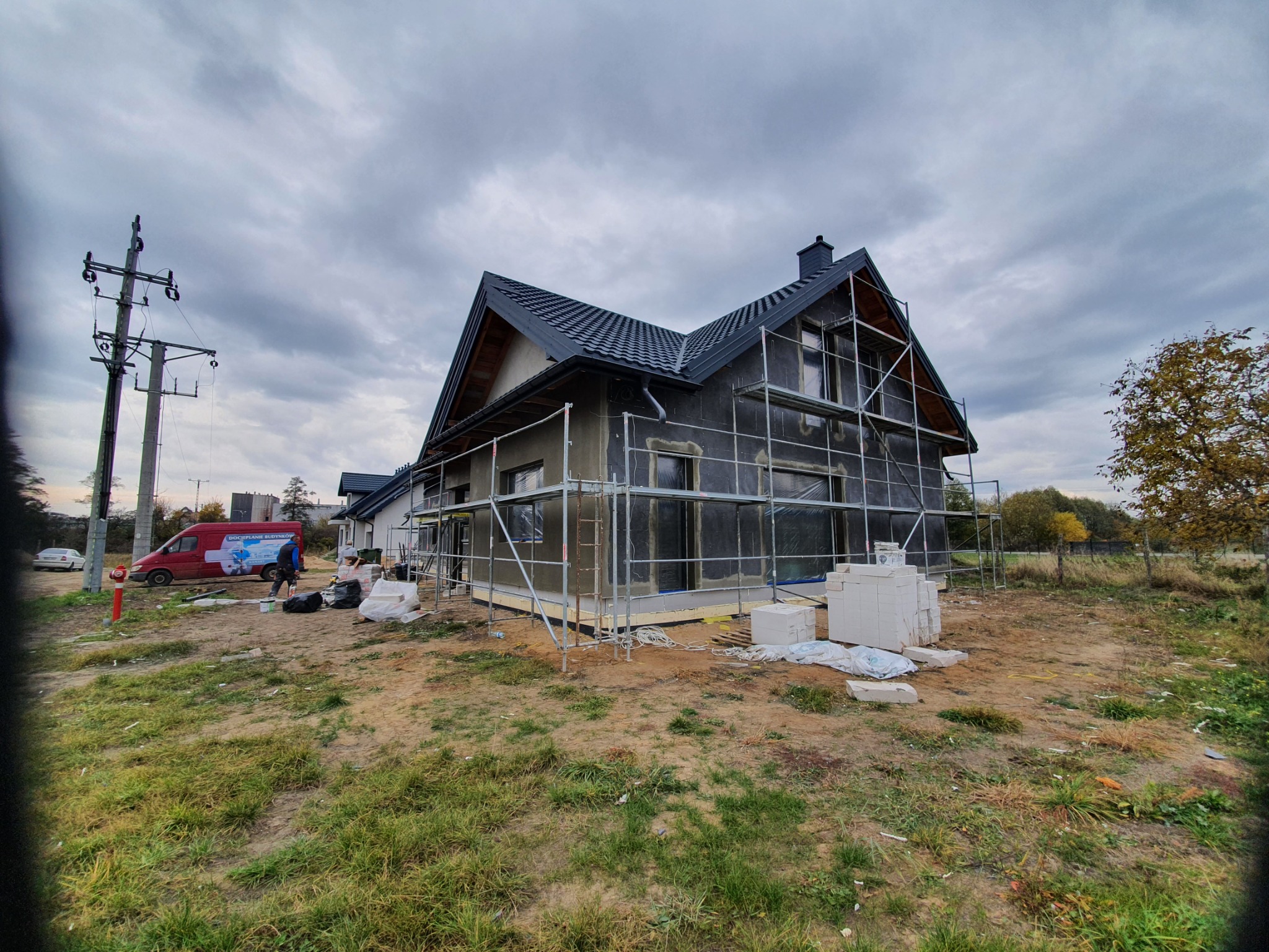 Dom w trakcie ocieplania elewacji, widoczne rusztowania, styropian i częściowo otynkowana ściana, na pierwszym planie porozrzucane materiały budowlane i trawa, w tle samochód dostawczy z reklamą...