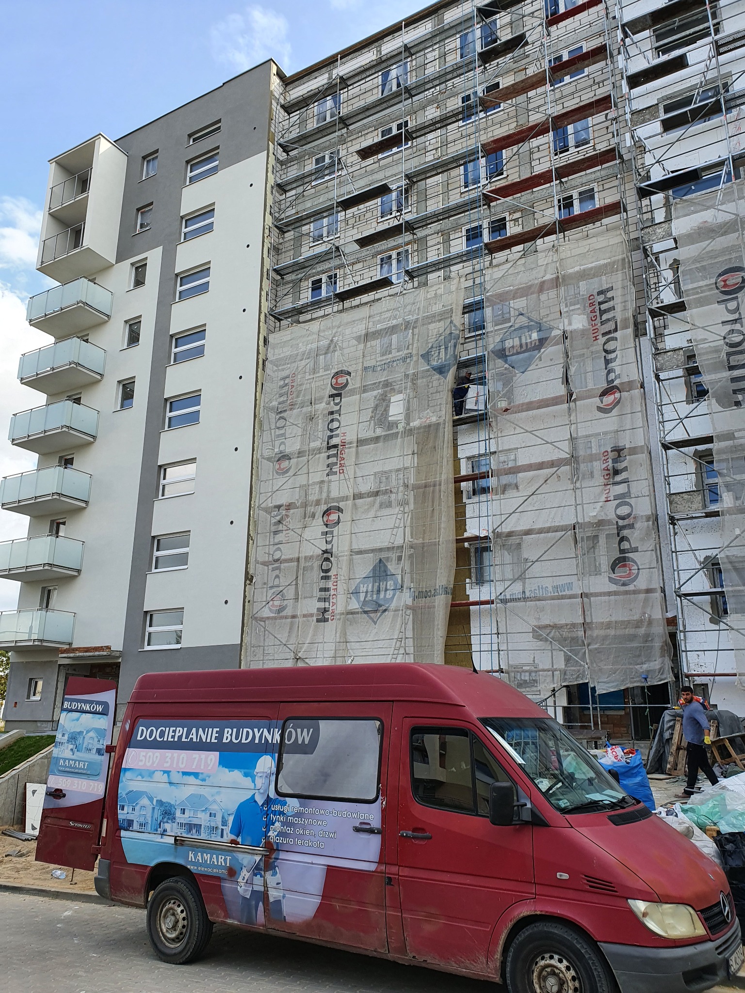Wielopiętrowy budynek w trakcie prac elewacyjnych, z widocznymi rusztowaniami i siatką ochronną, obok zaparkowany czerwony bus z reklamą firmy budowlanej.