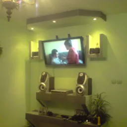 Ściana w odcieniach zieleni pokryta tapetą natryskową, z zamontowanym telewizorem, półkami i głośnikami, oświetlona wiszącym żyrandolem.