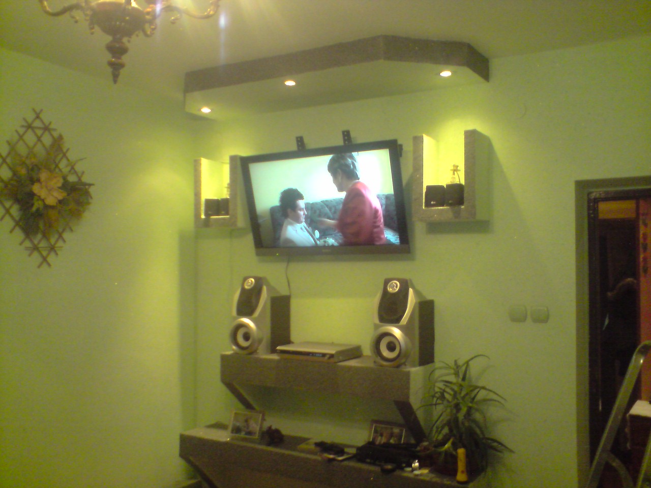 Ściana w odcieniach zieleni pokryta tapetą natryskową, z zamontowanym telewizorem, półkami i głośnikami, oświetlona wiszącym żyrandolem.