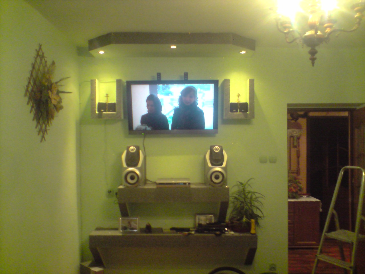 Zielone ściany w pokoju z telewizorem, głośnikami, półkami i dekoracyjną lampą, widoczne elementy tapety natryskowej.