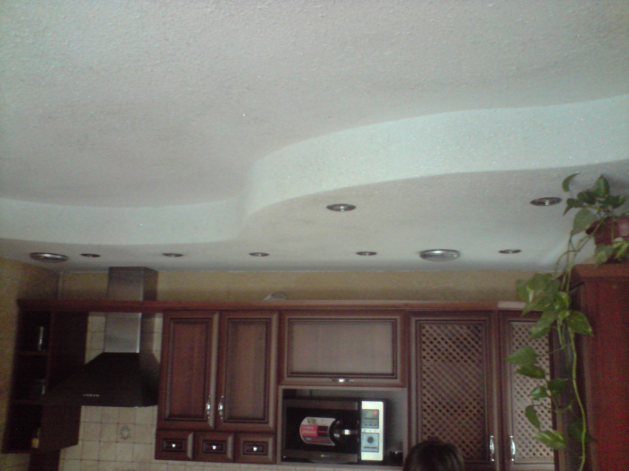 Biały, wielopoziomowy sufit z oświetleniem punktowym w kuchni z zabudową szafek wiszących i okapem.