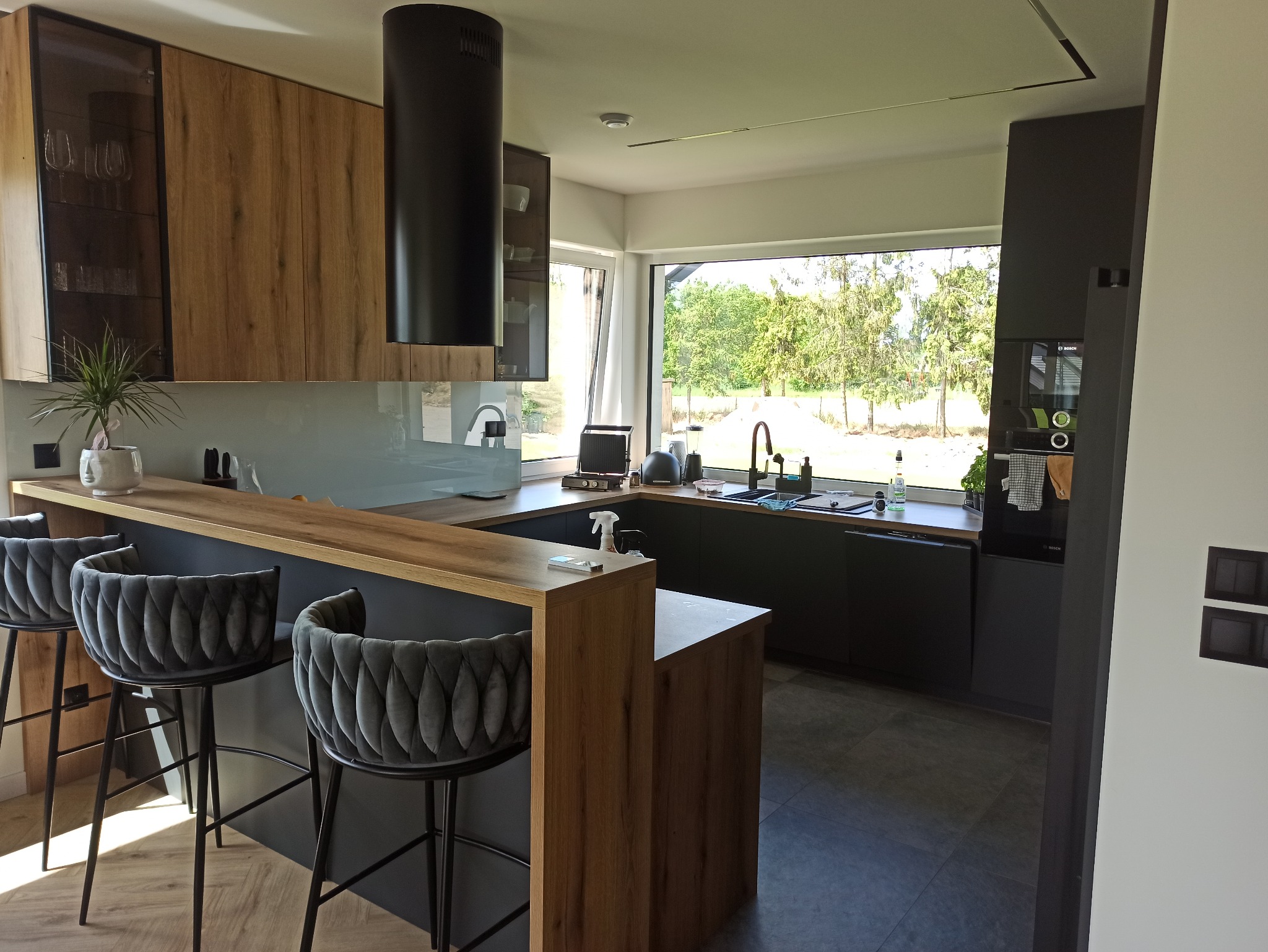 Nowoczesna kuchnia z drewnianymi i czarnymi elementami, wyspą i barowymi krzesłami. Duże okno z widokiem na zieleń. Minimalistyczny design.