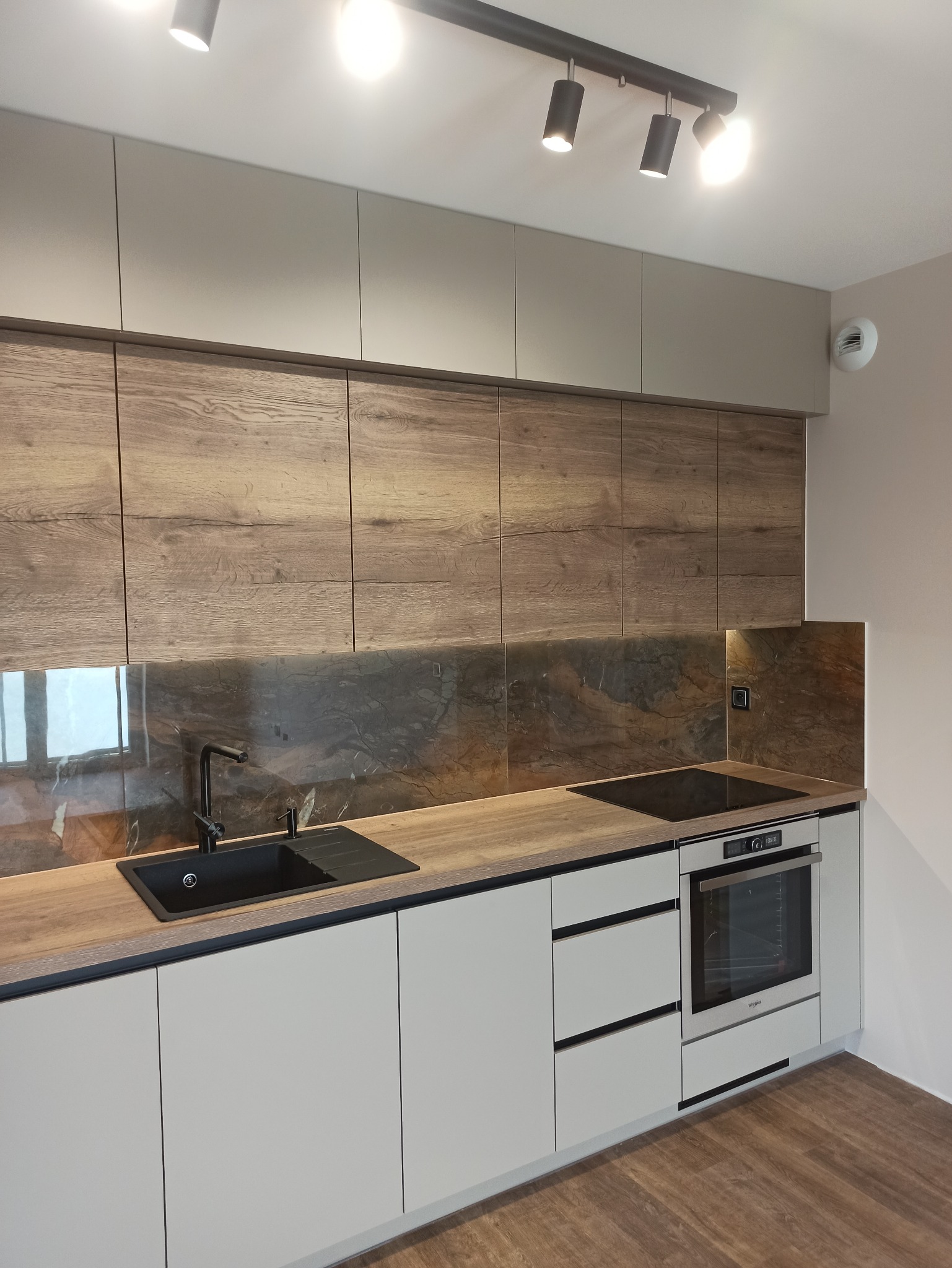 Nowoczesna kuchnia z drewnianymi szafkami górnymi i blatem, czarny zlew i bateria, białe szafki dolne z poziomymi uchwytami, oświetlenie punktowe.